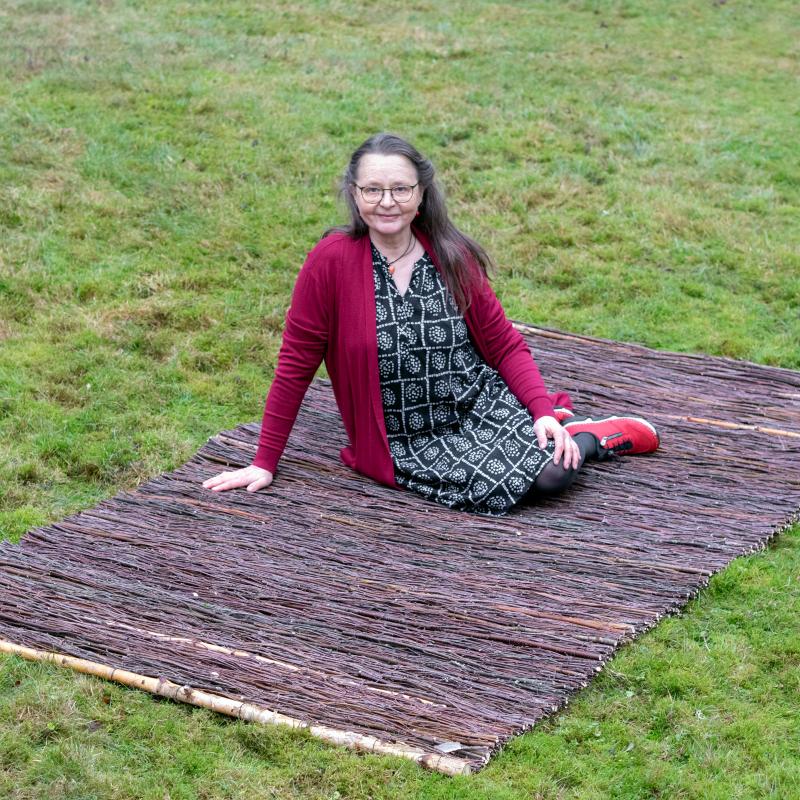 Tarja Heikkilä istuu risumaton päällä. Matto on näyttelyn "kynnysmatto".