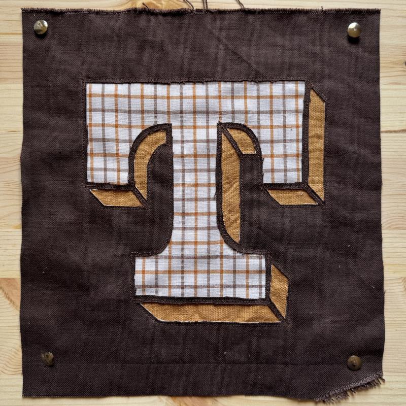 T (textil-lettering i återbruket material, 2025)